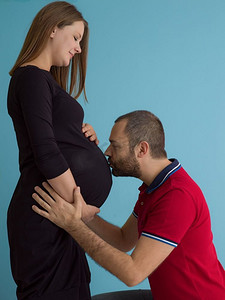 怀孕的夫妇摄影照片_怀孕的夫妇孤立在蓝色背景。一对快乐的年轻夫妇的肖像，男子拿着他的怀孕的妻子肚子隔绝在蓝色背景