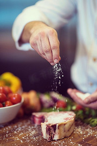 烘烤食物摄影照片_主厨把盐放在多汁的生牛排上，周围放着蔬菜放在木桌上。厨师把盐放在多汁的生牛排上