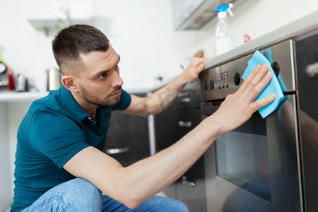 擦厨房摄影照片_家居和人的概念--男人擦桌子，用布清洗家里厨房的烤箱门。在家里厨房里拿着抹布清洁炉门的男人