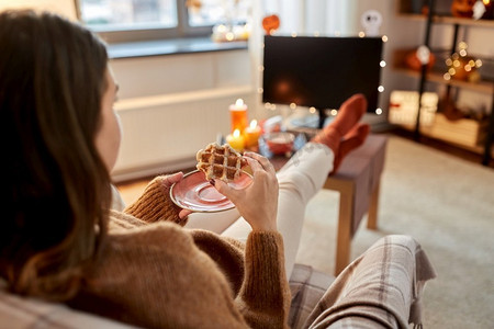 万圣节，假日和休闲的概念-年轻女子在舒适的家里看电视和吃华夫饼。万圣节前夕看电视吃华夫饼的女人