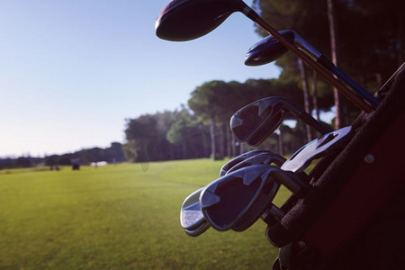高尔夫球杆图形摄影照片_在美丽的日出时分，高尔夫球包在球场上，球杆和球在前面。在球场上合上高尔夫球袋