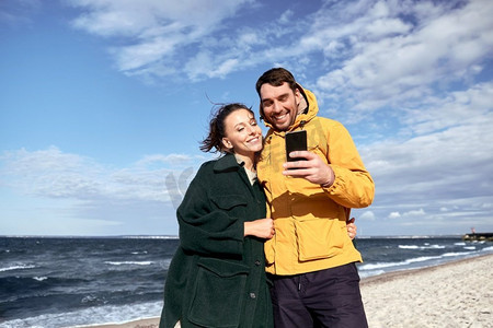 与人连接摄影照片_技术，关系和人的概念—快乐的夫妇与智能手机在秋天的海滩。夫妇与智能手机在秋季海滩