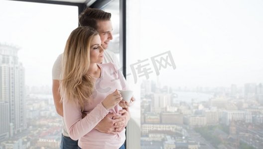 窗边浪漫摄影照片_在寒冷的冬日里，一对浪漫幸福的年轻夫妇在家中靠窗品尝早晨的咖啡。一对年轻夫妇在窗边喝着早晨的咖啡
