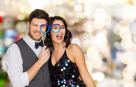 庆祝、娱乐和假期概念-幸福的情侣在灯光背景下戴着派对眼镜拍照。戴着派对眼镜的幸福情侣