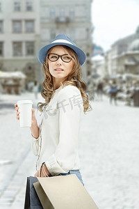 靠边摄影照片_年轻时髦的女人拿着购物袋，在购物后喝着咖啡，拿着一个咖啡外带杯子，在城市背景的映衬下站在一边。