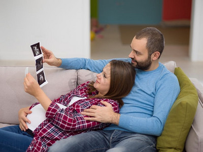 怀孕的夫妇摄影照片_怀孕的夫妇寻找婴儿的超声波。’年轻的怀孕夫妇寻找婴儿的超声波照片，而放松在沙发上在家里’