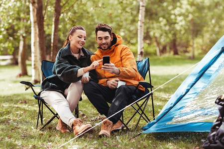 旅游工具摄影照片_露营，旅游和旅行概念—愉快的夫妇与智能手机喝茶在露营地。夫妇与智能手机喝茶在营地