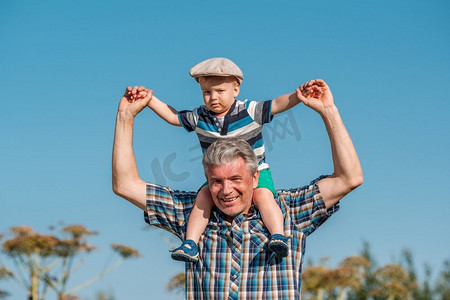 扛在肩上摄影照片_爷爷把蹒跚学步的孙子扛在肩上。在户外有骑在他的祖父的背上的孩子。 