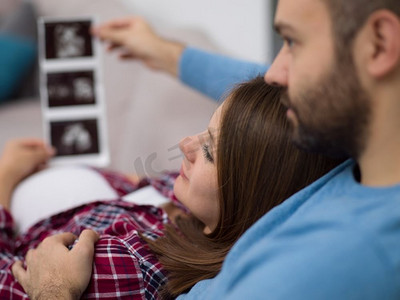 怀孕的夫妇看起来像婴儿&S；超声波。坐在家里的沙发上休息时，年轻的怀孕夫妇看起来像婴儿和S的超音波照片