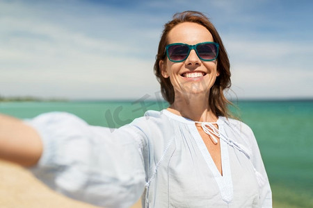 暑假休闲概念--戴着墨镜的快乐微笑的女人在海滩自拍。一名戴墨镜的女子在夏日海滩自拍