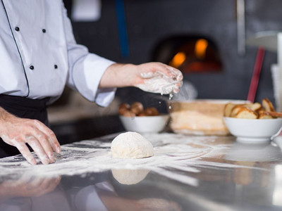 意大利面意大利面摄影照片_厨师把面粉洒在厨房桌子上的新鲜披萨面团上。厨师在新鲜的披萨面团上撒面粉