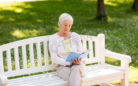 老年、退休与人的概念--坐在夏令营长椅上看书的资深女性。老年妇女在夏季公园看书