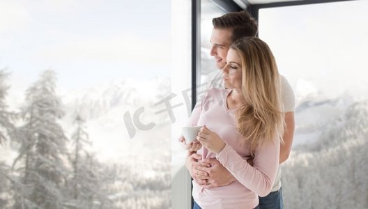 在寒冷的冬日里，一对浪漫幸福的年轻夫妇在家中靠窗品尝早晨的咖啡。一对年轻夫妇在窗边喝着早晨的咖啡