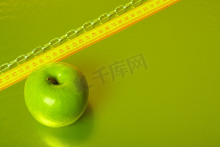 链子摄影照片_绿色的苹果放在金色的背景上，上面有卷尺和链子。健康的生活方式--营养与锻炼