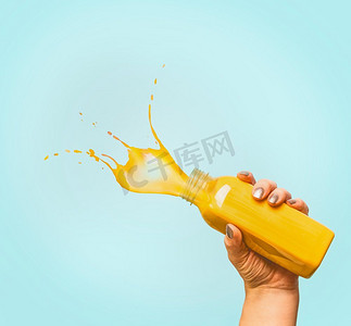 水果喷溅摄影照片_女性手持瓶，黄色喷溅夏季饮料：冰沙或果汁，蓝色背景，复制空间