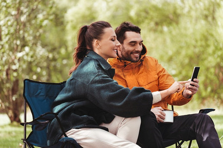 旅游小程序摄影照片_露营、旅游和旅行概念-在露营地点与智能手机喝茶，这是一对幸福的情侣。与智能手机一起在露营地喝茶