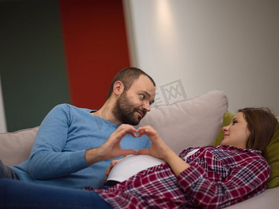 示爱男人摄影照片_男人和孕妇显示心脏症状。快乐的男人和孕妇显示心脏标志与手指，而在家里的沙发上放松
