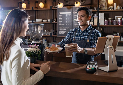 食堂外卖摄影照片_亚洲咖啡师的肖像举行带走咖啡和羊角面包杯和服务给顾客在咖啡馆与其他咖啡师工作在背景