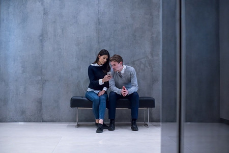 一对年轻的商业夫妇使用智能手机，而坐在办公室大厅的长凳上休息