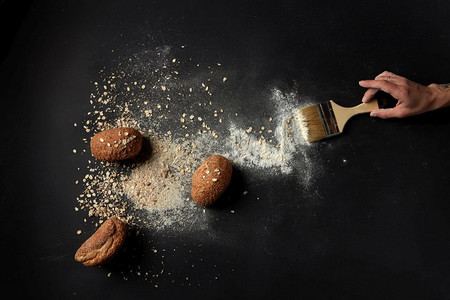 面包的顶视图与燕麦片和面粉，人的手拿着刷子在黑色背景.新鲜烤面包刷
