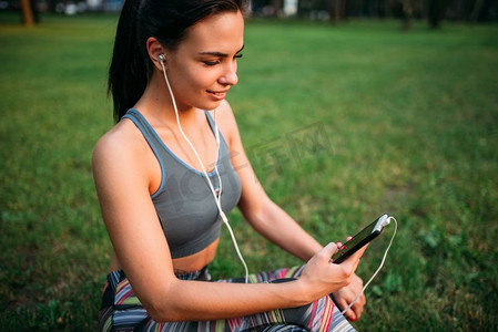 戴着耳机的苗条女人坐在草地上放松。运动女孩在户外健身锻炼