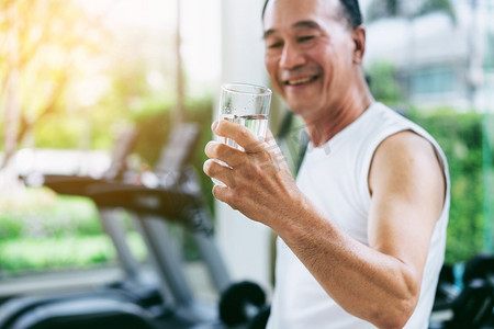 老年人喝酒摄影照片_老年人运动后在健身房健身中心喝矿泉水。老年人健康的生活方式。