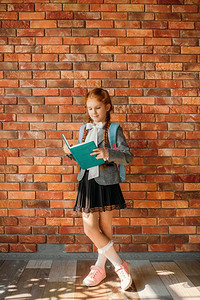 有书包的可爱的女学生读教科书，在背景的砖墙。可爱的女学生背包和书姿势在学校