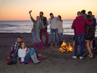 年轻夫妇坐在周围篝火在海滩上的朋友在日落喝啤酒。夫妇享受与朋友在日落在海滩