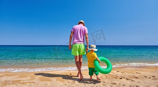 两岁的小男孩和父亲一起在海滩上散步，手里拿着充气戒指。暑假全家度假。希腊西索尼亚。