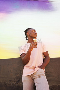 一个年轻英俊的黑人男子穿着粉红色的T恤，在夏天的日出或晴天的墙上拿着一个冰淇淋吃