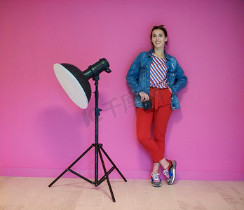 女摄影师拿着相机在她的手和靠在粉红色的墙壁。摄影棚内的专业摄影设备