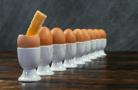 第一食品摄影照片_在一张木桌上的白色蛋杯里的煮鸡蛋排第一个蛋被打破了与一个烤面包士兵浸在它