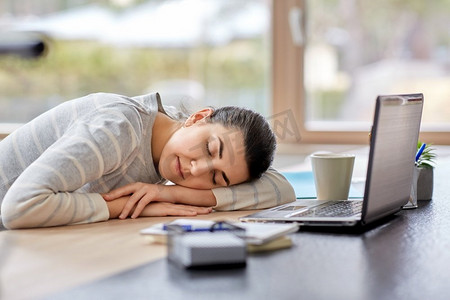 人和生活方式的概念—疲倦的妇女睡在桌上与膝上型计算机在家庭办公室。疲惫的女人睡在桌子上与笔记本电脑在家里