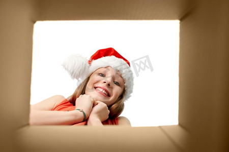 圣诞老人帽子摄影照片_圣诞节，礼物和惊喜概念-戴着圣诞老人帽子的惊喜女孩看着礼品盒。惊讶的女孩在看圣诞礼品盒
