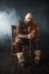 穿着北欧传统服装坐在椅子上的有胡子的维京人手持斧头，野蛮人的形象。古代战士在烟雾在黑暗的背景