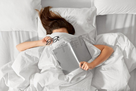人，睡觉时间和休息的概念--躺在床上的女人，盖着白色的毯子或羽绒被，带着书和眼镜。躺在床上的女人，拿着书和眼镜