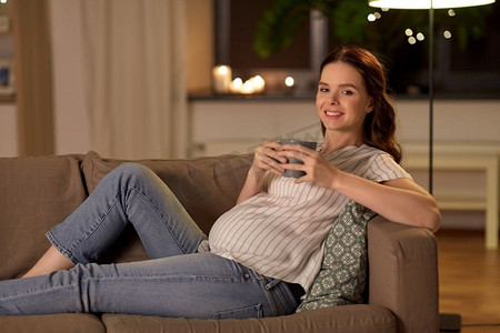 怀孕和人的概念—愉快的微笑孕妇喝茶在家里的沙发上。微笑的孕妇喝茶在家