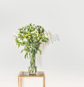 美丽的绿色花束与落花瓣在玻璃花瓶在白色墙壁背景。家居内饰和装饰理念
