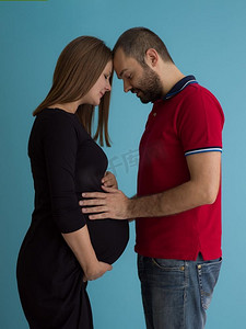 怀孕的夫妇摄影照片_怀孕的夫妇孤立在蓝色背景。一对快乐的年轻夫妇的肖像，男子拿着他的怀孕的妻子肚子隔绝在蓝色背景