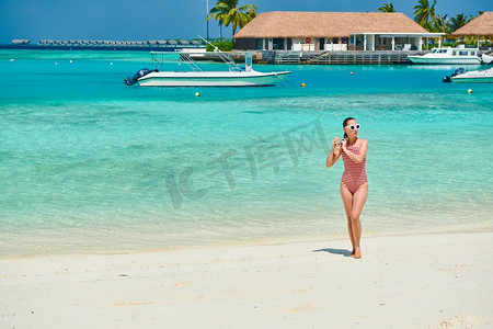 连体比基尼摄影照片_穿着连体式泳衣的女子在白色沙滩上行走。在马尔代夫度暑假。