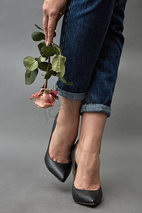 情人玫瑰摄影照片_一个女孩在牛仔裤和黑色的鞋子持有粉红色的玫瑰媒体在灰色背景。女孩拿着一个粉红色的玫瑰媒体