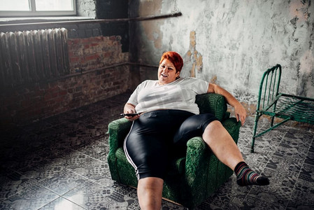 超重的女人坐在椅子上看电视。不健康的生活方式肥胖