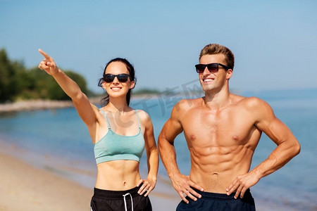 健身，运动和生活方式的概念—快乐的夫妇在运动服和太阳镜在海滩。快乐的夫妇在运动衣服和阴影在海滩上