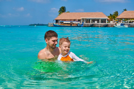 全家一起去旅行摄影照片_三岁的学步男孩和父亲一起学习游泳。暑假全家去马尔代夫度假。