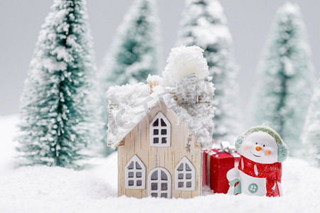 冬天的雪人摄影照片_小装饰雪人与礼物在冷杉林附近的木屋在落雪。冬天的雪人和房子