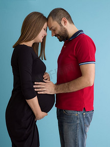 怀孕的夫妇孤立在蓝色背景。一对快乐的年轻夫妇的肖像，男子拿着他的怀孕的妻子肚子隔绝在蓝色背景