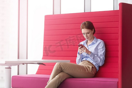 年轻女商人坐在办公室的红色座椅上用智能手机发信息