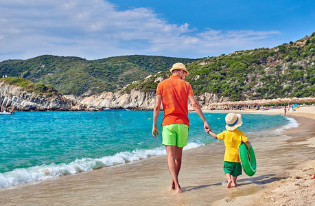 两岁的小男孩和父亲一起在海滩上散步，手里拿着充气戒指。暑假全家度假。希腊西索尼亚。