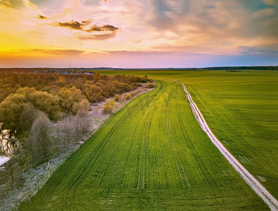 春路摄影照片_乡村土路穿春绿田。傍晚的乡村风景。四月日落在白俄罗斯鸟瞰图