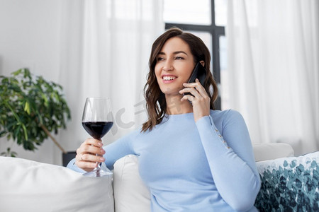 休闲和人的概念—快乐的女人打电话在智能手机和喝红葡萄酒在家里。女人打电话给智能手机喝红酒